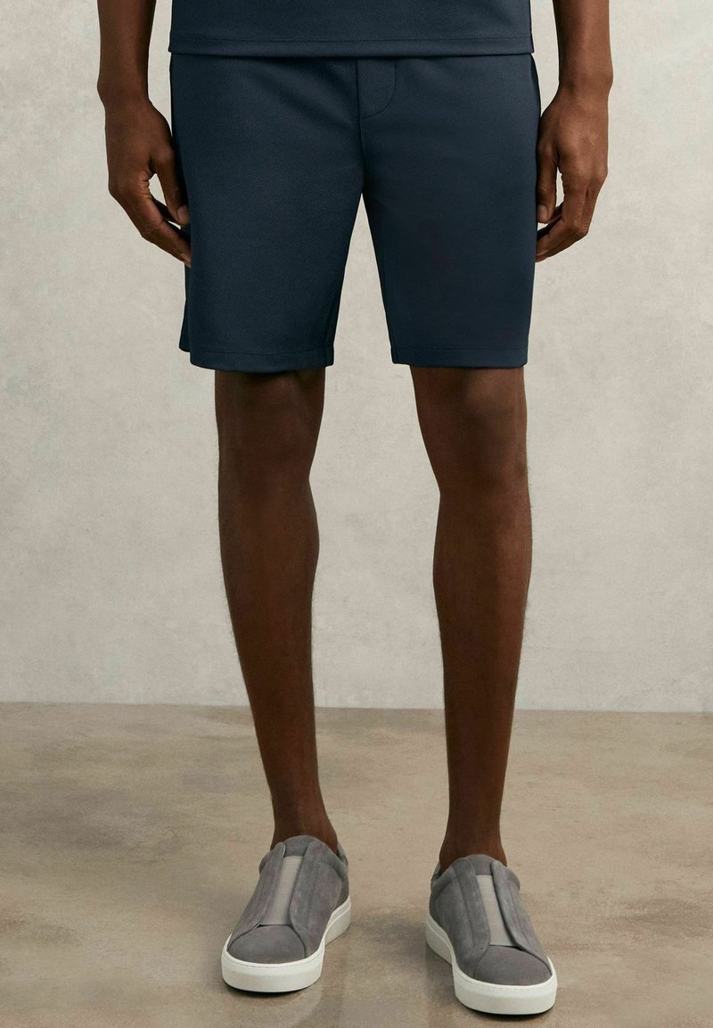 Marineblaue Shorts aus strukturiertem Stoff, mit elastischem Bund und modernem Schnitt, kombiniert mit grauen Slippers.