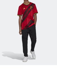 Maglia da calcio rossa con strisce diagonali nere, bordature gialle, logo sul petto, abbinata a pantaloni neri e scarpe da ginnastica bianche.