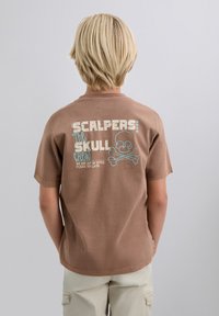 Niño rubio con camiseta marrón que tiene un gráfico de calavera azul y blanca y el texto "Scalpers The Skull Crew, Estamos fuera de la oficina, por favor inténtalo más tarde."