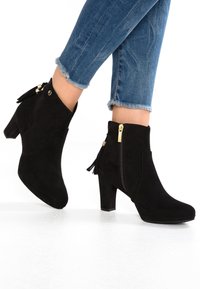 Černé nubukové kotníkové boty s masivním podpatkem, postranním zipem a akcentem v podobě třásní. K botám jsou nošeny modré džíny s roztrhaným lemem.