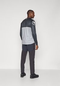 Langärmliges graues und schwarzes Sportoberteil mit Schulterlogo, kombiniert mit dunklen Hosen mit seitlichem Reißverschluss. Beide sind aus glattem Stoff gefertigt.