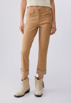 Persona che indossa pantaloni color cammello risvoltati con polsini decorati e stivaletti beige con tacco basso.