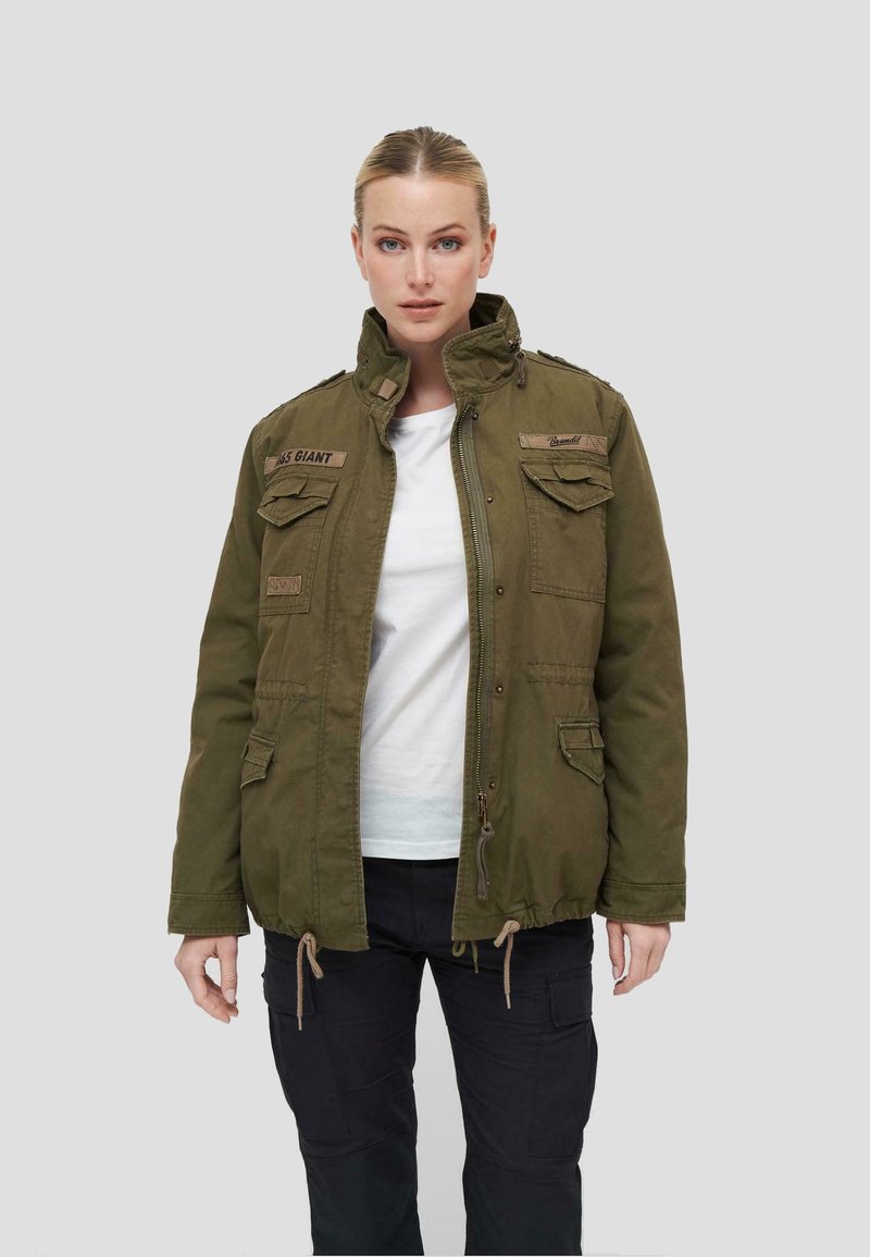Chaqueta militar de color verde olivo con cuello alzado, dos bolsillos en el pecho y cintura ajustable con cordón. Presenta detalles de costura en los hombros y acentos de parches.