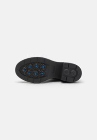 Suela de zapato de goma negra con múltiples patrones de agarre circulares y acentos azules. Presenta un tacón plano y una superficie texturizada con ranuras definidas.