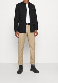 Camicia nera a maniche lunghe con chiusura a bottoni, indossata sopra una t-shirt bianca. Abbinata a pantaloni beige e stivali neri alla caviglia.