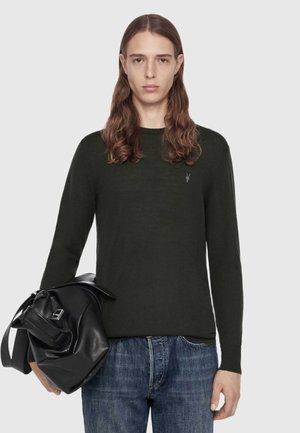 Ung person med langt brunt hår iført en mørkegrøn sweater og blå jeans, holder en sort lædertaske mod en ensfarvet baggrund.