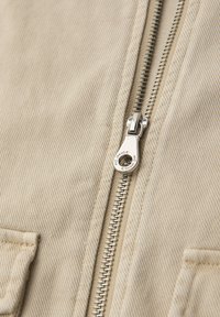 Tela beige con una cremallera metálica parcialmente cerrada, mostrando los dientes detallados de la cremallera y la lengüeta de tirador sobre una prenda texturizada.