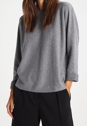 Kobieta w szarej, długorękawowej, teksturowanej bluzce z podwiniętymi mankietami i luźnych czarnych spodniach, stojąca z jedną ręką w kieszeni.