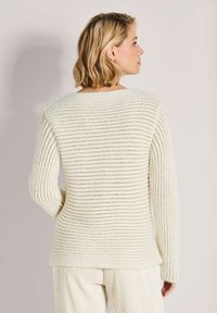 Frau mit kurzem blonden Haar, die einen cremefarbenen, gerippten Strickpulli und passende Hose trägt, von hinten vor einem schlichten Hintergrund gesehen.