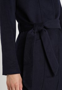Marineblauer Wickelmantel aus dickem Stoff mit einem gebundenen Gürtel in der Taille, langen Ärmeln und seitlichen Taschen mit glatter Textur.