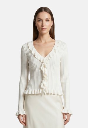 Donna con capelli castani lisci che indossa un cardigan bianco a coste con volant lungo la scollatura, davanti, ai polsini e all'orlo, rivolta verso il davanti.