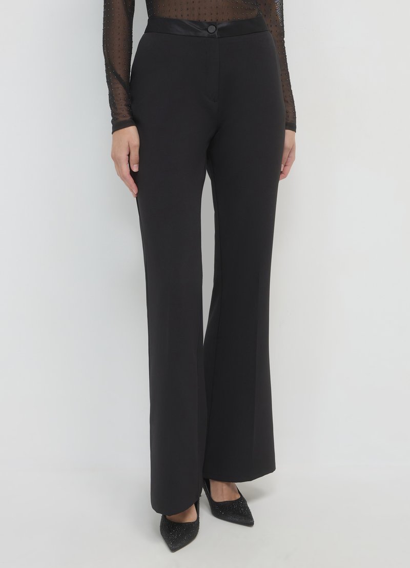 Pantalon évasé noir à la texture lisse, taille en satin, fermeture avant à boutons, coupe ajustée, associé à des talons noirs pointus.