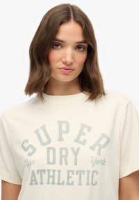 Beige t-shirt lavet af bomuld. Har fed grå tekst i en bue: "SUPER DRY ATHLETIC" og lille "New York" nedenunder. Korte ærmer.