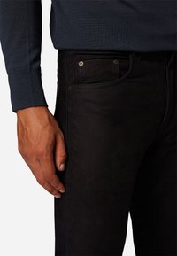 Zwarte, slim-fit spijkerbroek met een gladde textuur, voorzien van goudkleurige hardware en frontzakken. Draagt een donkerblauwe trui.