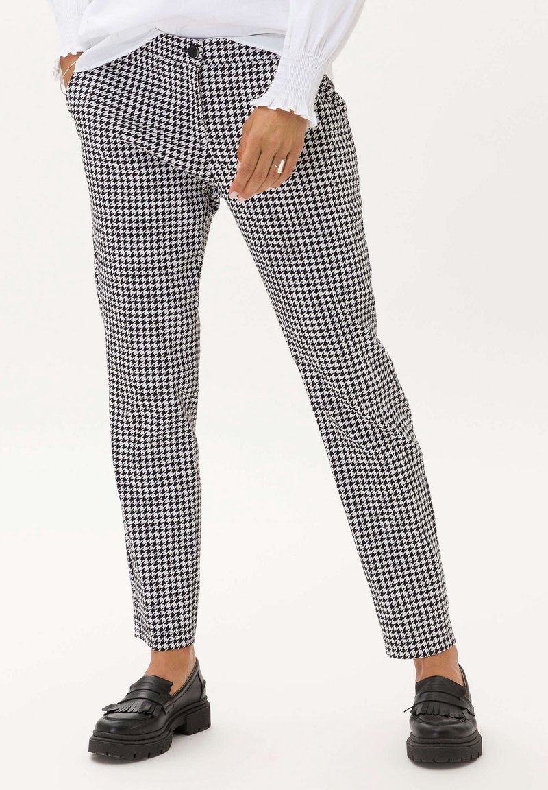Femme portant un pantalon slim noir et blanc à motif pied-de-poule, un chemisier blanc avec des poignets à volants, et des mocassins en cuir noir avec un détail à franges.