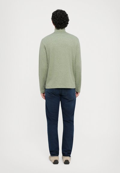 Πράσινο μακρυμάνικο πουκάμισο με ψευδές λαιμό, συνδυασμένο με σκούρα μπλε τζιν και ανοιχτόχρωπα παπούτσια, εμφανίζεται από πίσω σε έναν απλό φόντο.