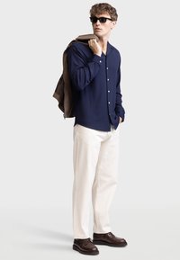Navy button-up shirt with ribbed texture, paired with cream chinos and dark brown lace-up shoes; model holds a brown jacket over one shoulder.