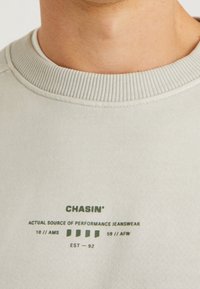 Sudadera de color gris claro con cuello acanalado, que presenta texto verde: "CHASIN' Actual Source of Performance Jeanswear." Textura suave, diseño casual.