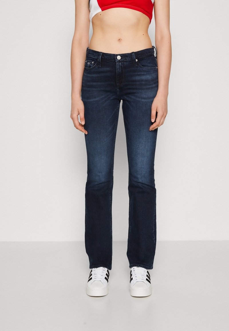 Personne portant un jean bleu foncé à coupe droite et des baskets blanches avec des bandes noires, debout devant un fond uni.