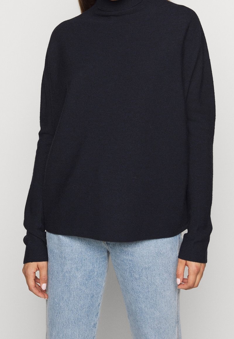 Femme portant un pull à col roulé noir à manches longues et un jean bleu clair, debout devant un fond clair uni.