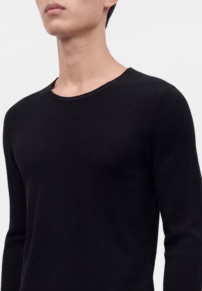 Homme portant un pull noir uni à manches longues avec un col rond sur un fond gris clair.