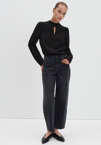 Donna che indossa un top lungo nero con un'apertura frontale, jeans neri a vita alta sbiaditi e ballerine nere a punta, in piedi contro uno sfondo semplice.