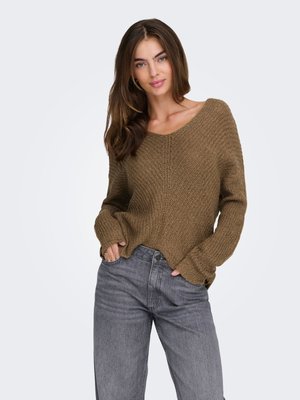 Kvinde med langt brunt hår iført en brun strikket sweater og grå jeans, poserer med hænderne i lommerne mod en ensfarvet baggrund.
