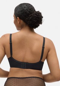 Sujetador de tela negra con tirantes ajustables, cierre de gancho y ojo en la parte posterior y una textura suave. Tela de lunares transparentes visible por debajo.