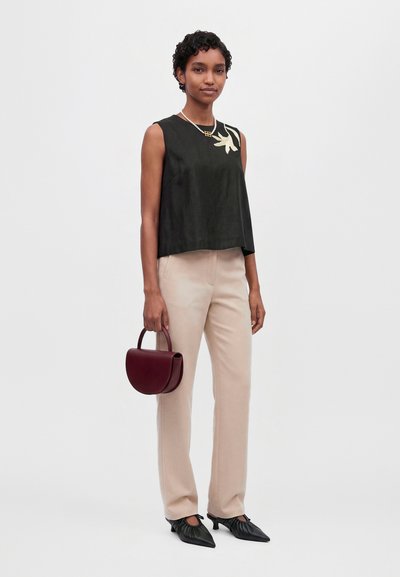 Un mannequin porte un haut noir sans manches à motif floral, un pantalon crème et des ballerines noires. Elle tient un sac à main bordeaux devant un fond uni.