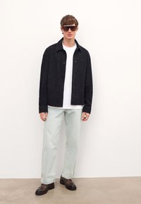 Giacca in suede nera con chiusura a bottone, abbinata a una maglietta bianca, jeans azzurri chiari e scarpe marroni, indossata con occhiali da sole scuri.
