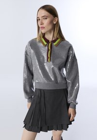 Grijze sequined pullover met een levendige gestreepte kraag in rood en groen, gecombineerd met een geplooide zwarte rok met een aansluitende tailleband.