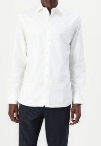 Vit långärmad skjorta med knäppkrage och framknäppning. Tillverkad av ett slitstarkt tyg; har en skräddarsydd passform och en minimalistisk design.