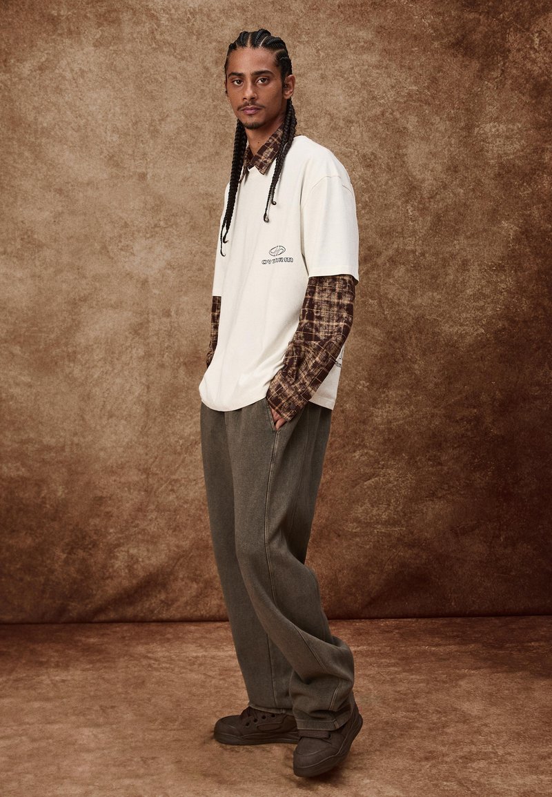 Bijela oversized majica preko smeđe karirane košulje dugih rukava, uparena sa sivim širokim hlačama i crnim tenisicama. Teksturalna pozadina.