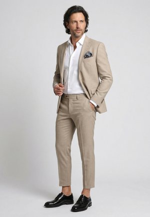 Mand iført beige jakkesæt med hvid skjorte, paisley lommetørklæde og sorte sko, poserer mod en ensfarvet lys grå baggrund.