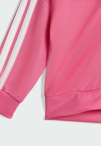 Różowa sportowa bluzka z długim rękawem z białymi paskami, teksturowana tkanina i prążkowane mankiety. Design podkreśla sportowy styl.