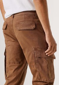 Bruine cargobroek gemaakt van duurzaam stof, met zijzakken en achterflappen, een ontspannen pasvorm en zichtbare stiksel details.