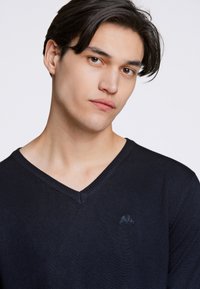 Marineblauer V-Ausschnitt-Pullover in glatter Stricktextur mit dezentem Logo-Stick auf der Brust, der eine taillierte Silhouette und ein klares Design zeigt.