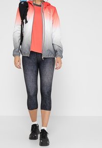 Lehká gradient bunda od červené po šedou, kombinovaná s tmavými vzorovanými capri legínami a černými teniskami. Bunda má kapuci a zip.