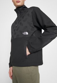 Svart fleece-pullover med halvmatchande krage, logotyp på bröstet och ett strukturerat mönster. Inkluderar en framficka och elastiska ärmslut.