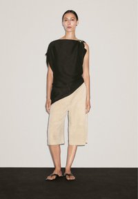 Top noir asymétrique avec un design drapé, assorti à un pantalon beige court. Accentué par des éléments en métal sur l'épaule. Sandales plates.