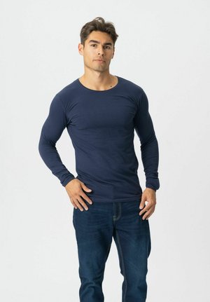 Bărbat în picioare pe fundal alb, purtând o cămașă cu mâneci lungi bleumarin și blugi de culoare albastru închis, cu o mână pe șold și cu o expresie neutră.