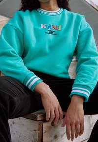 Persona seduta su una panchina di legno che indossa una felpa turchese con il logo "KANI" e pantaloni neri, mani appoggiate sulle ginocchia.