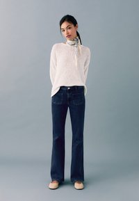 Kevyt, teksturoitu valkoinen neule yhdistettynä tummansinisiin leveälahkeisiin farkkuihin ja beigekiilakekkereihin; tyylitelty kuviollisella kaulahuivilla.