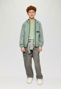 Lichtgroene waterdichte jas met een capuchon, zwarte rits, gedragen over een bijpassend shirt. Grijze cargobroek met meerdere zakken en witte sneakers.