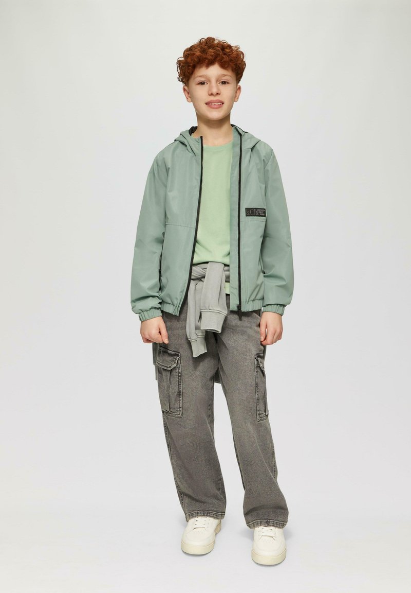 Lichtgroene waterdichte jas met een capuchon, zwarte rits, gedragen over een bijpassend shirt. Grijze cargobroek met meerdere zakken en witte sneakers.