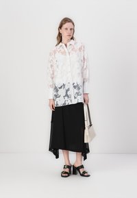 Hvid bluse med blomsterdesign i gennemsigtigt materiale, lange ærmer, knaplukning foran, samt sort maxi nederdel, parret med sorte hæl sandaler og en beige taske.