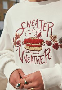 Biała bluza z haftem "Sweater Weather". Centrum ozdobione postacią Snoopy'ego w czerwonej i beżowej bluzie, otoczone liśćmi i żołędziami.