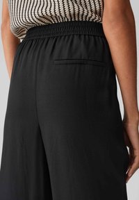 Schwarze weit geschnittene Hose hat einen elastischen Bund und eine kleine Gesäßtasche. Der Stoff hat eine glatte Textur mit subtilen Falten.