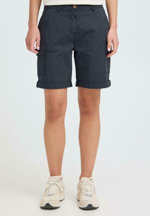 Shorts cargo bleu marine à revers roulé portés par une personne debout, associés à des baskets blanches, sur un fond clair uni.