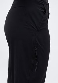 Κοντινό πλάνο μαύρου εξωτερικού παντελονιού με τσέπη με φερμουάρ και λογότυπο Salomon στο μηρό, με απλό ανοιχτό φόντο.